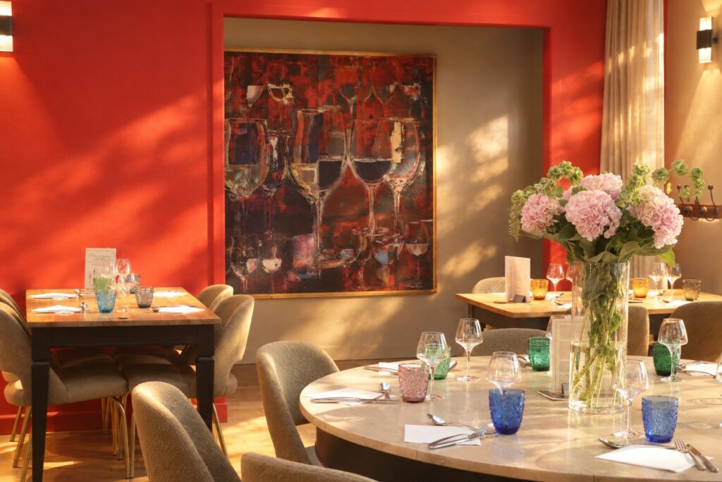 L'Entre-Soi : mur rouge, grand tableau de verres de vin, table ronde dressée et bouquet d'hortensias dans la lumière du soir